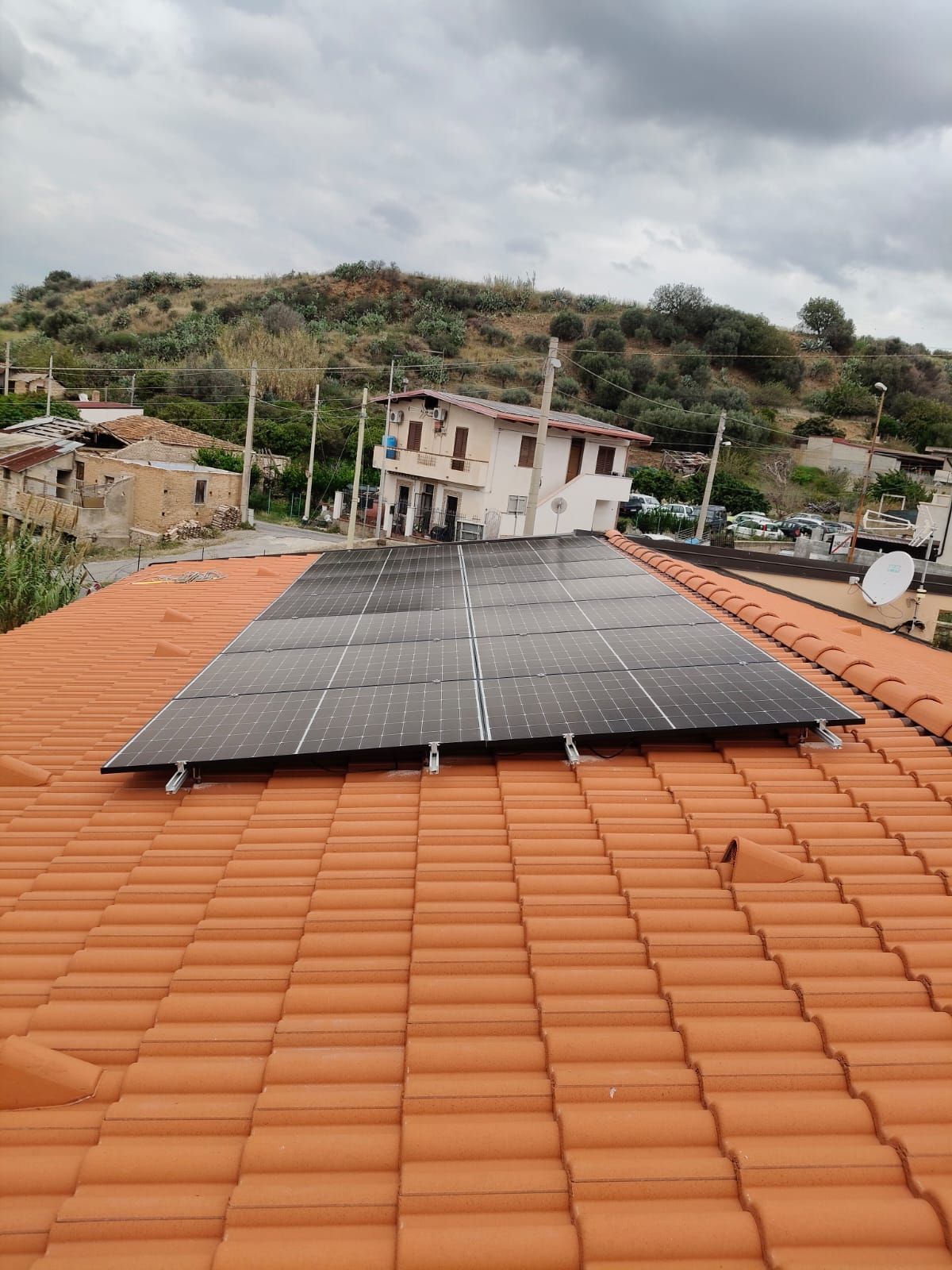 Pannelli solari sul tetto di una casa con tegole rosse, con colline e cielo nuvoloso sullo sfondo.