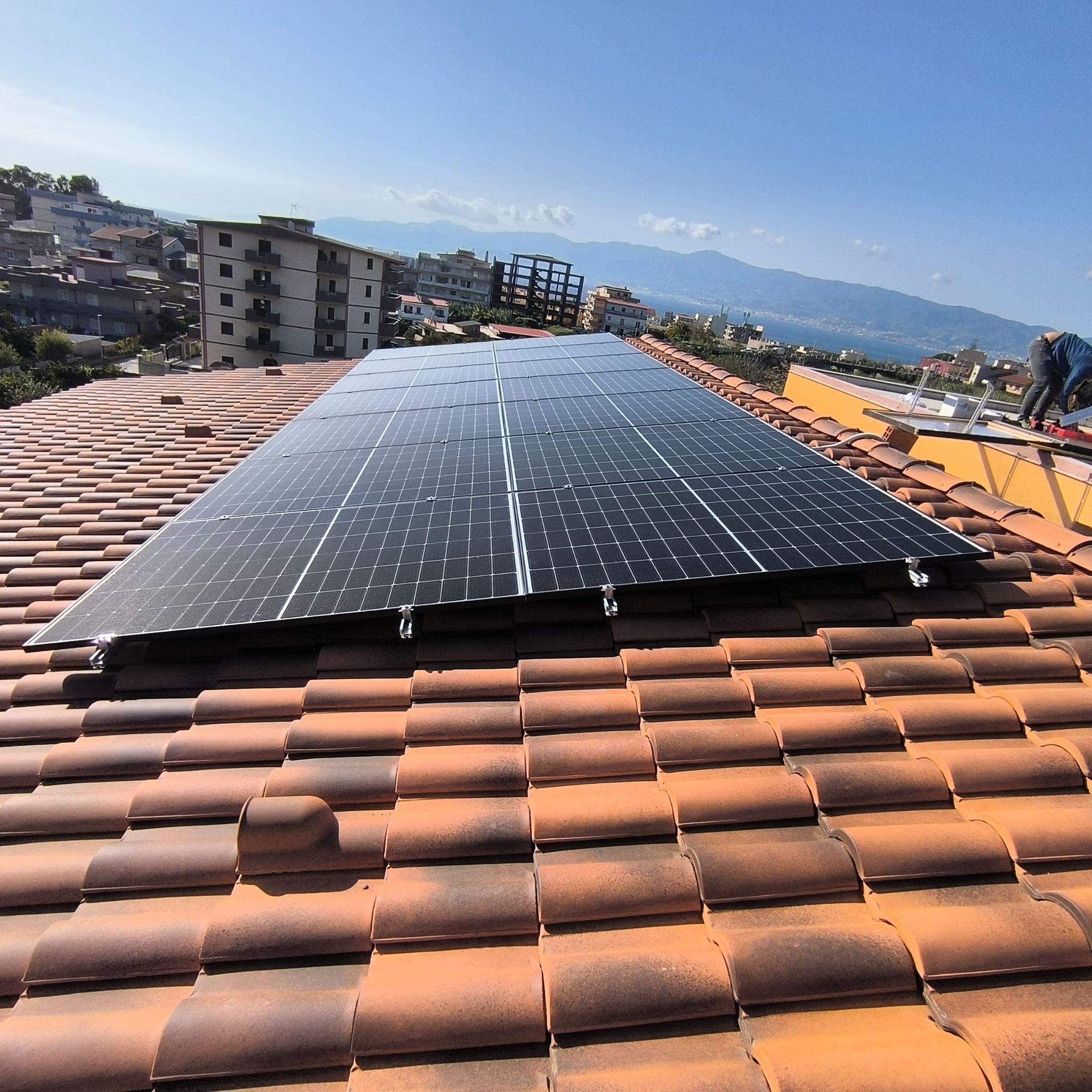 Pannelli solari installati su un tetto di tegole in terracotta, con panorama urbano e montagne sullo sfondo.