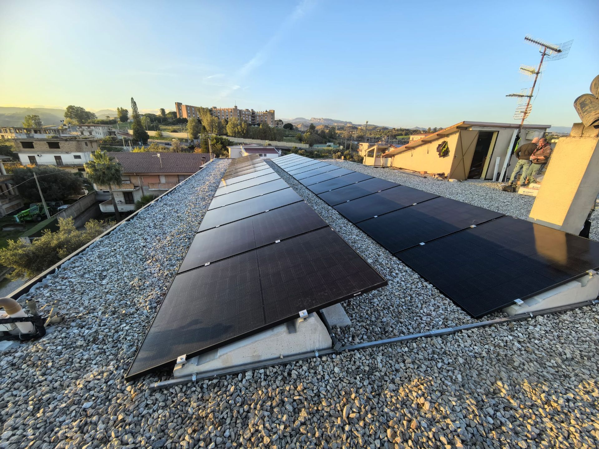 Pannelli solari installati su un tetto di ghiaia al tramonto, con gli edifici della città sullo sfondo.