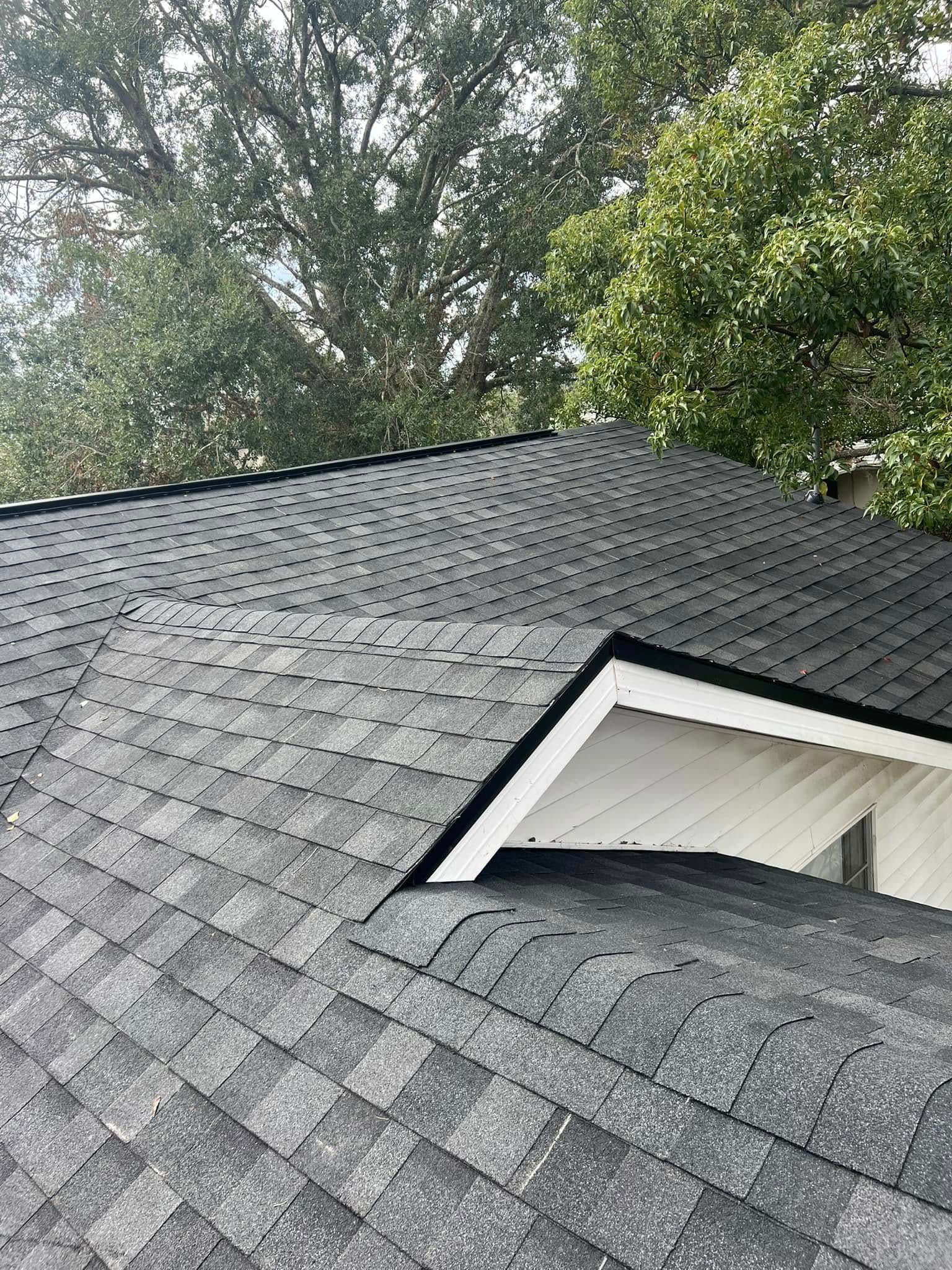 Damaged gray shingle roof with a hole exposing white attic framing and trees in the background