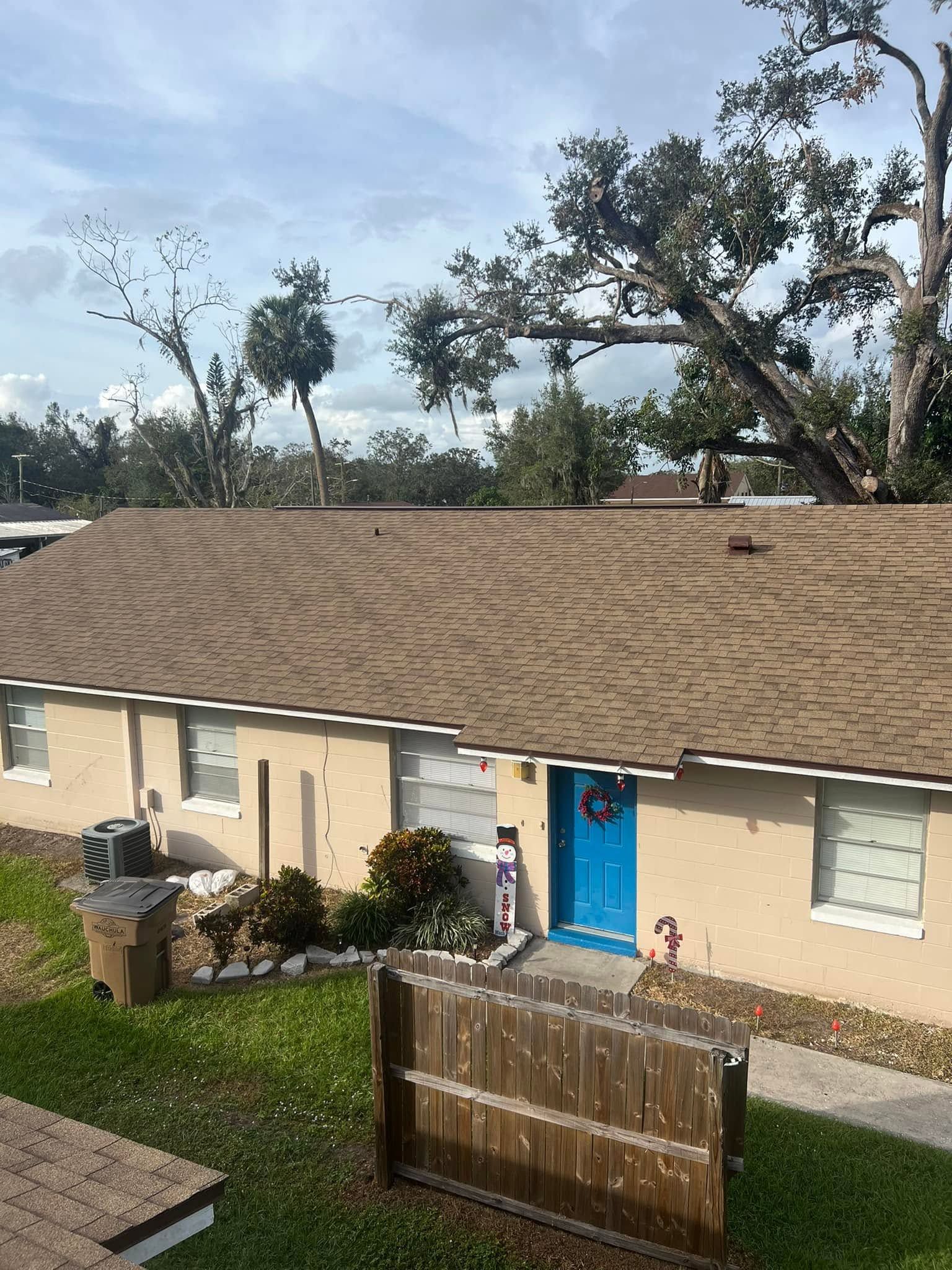 Single-story beige house with blue door, fenced yard, and fallen tree branches on the roof after a storm