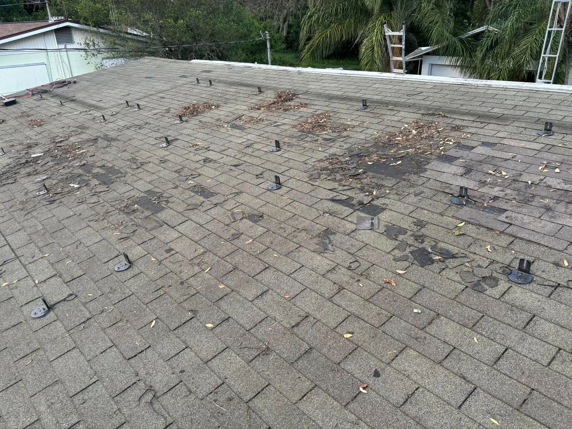 Damaged shingled roof with debris and a ladder near the edge, surrounded by trees and a nearby building.