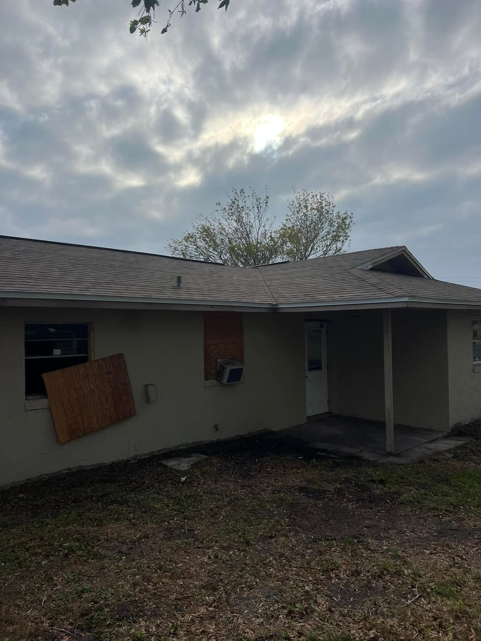 Damaged house with boarded window, sagging roof, and debris-strewn yard under a cloudy sky