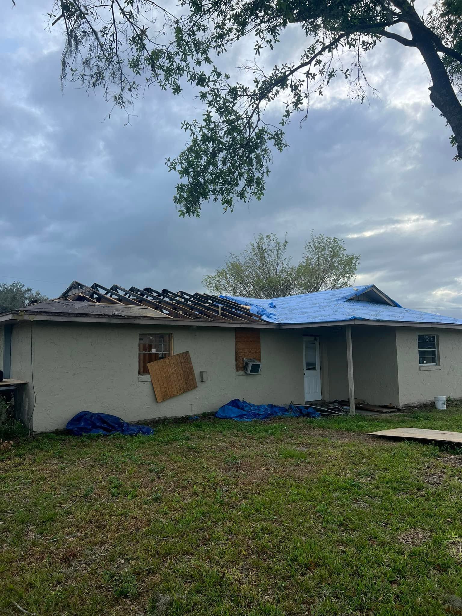 Single-story house with storm-damaged roof covered by blue tarp under cloudy sky