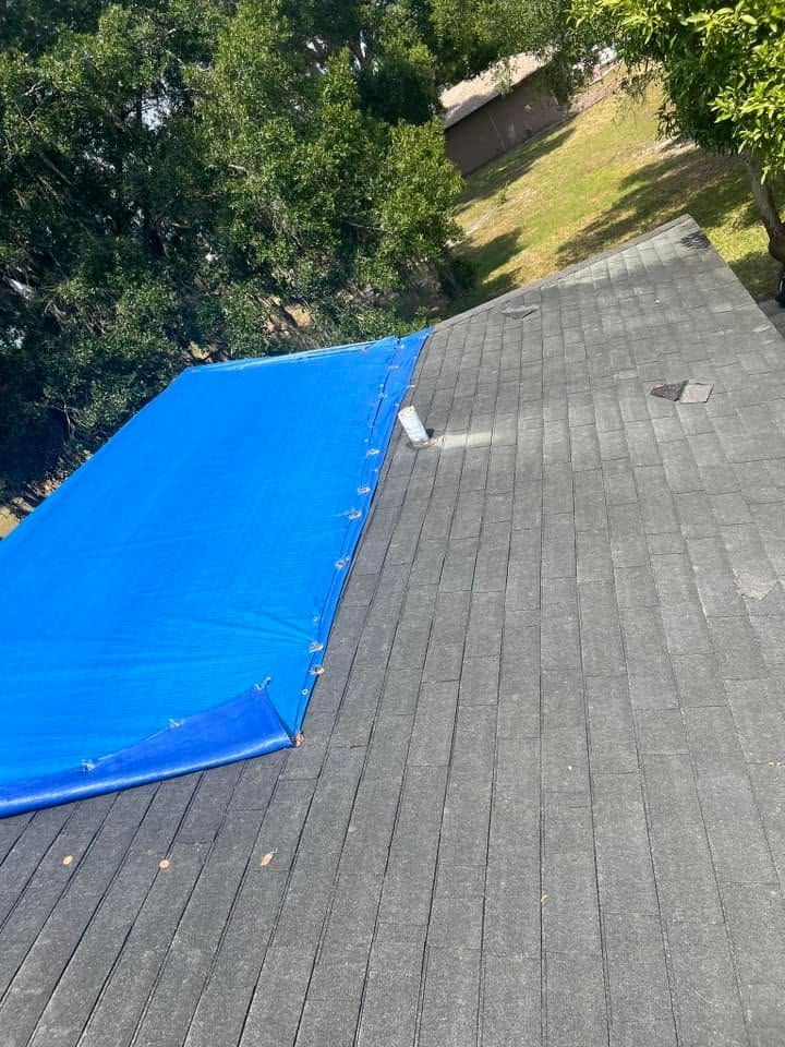 Rooftop with gray shingles and a bright blue tarp covering part of the left side, surrounded by trees.