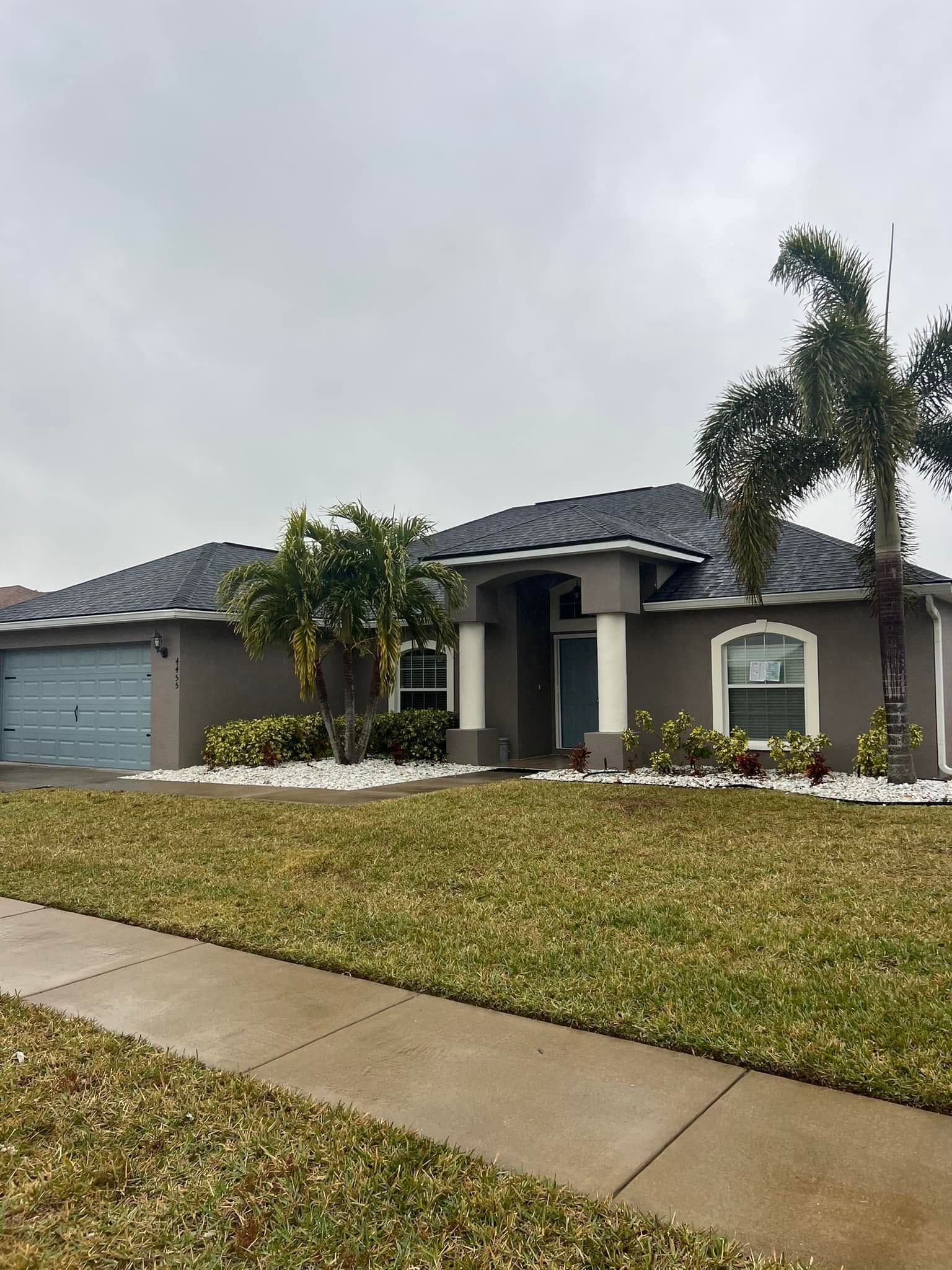 Single-story gray house with a tiled roof, front lawn, palm tree, and driveway under a cloudy sky
