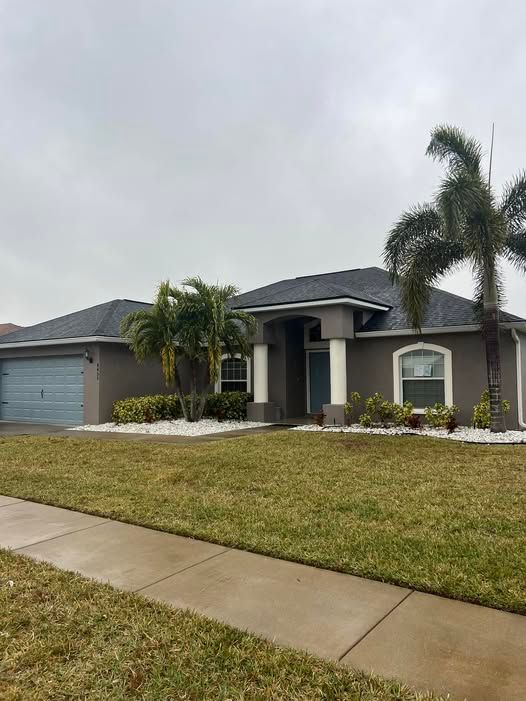 Suburban single-story house with gray roof, palm trees, and a wet sidewalk under cloudy skies