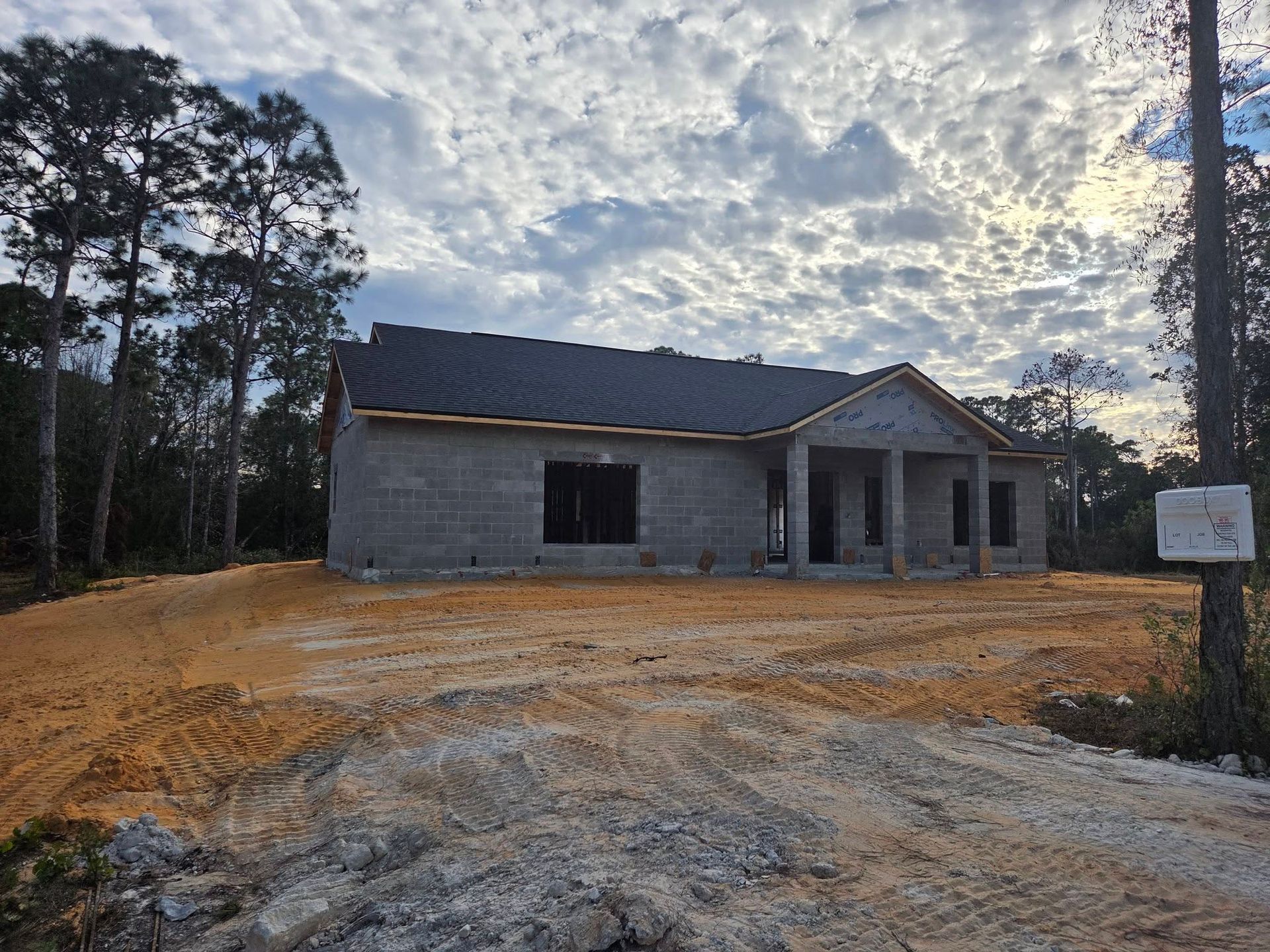 Unfinished gray cinderblock house under construction on a dirt lot at sunset, with trees nearby.