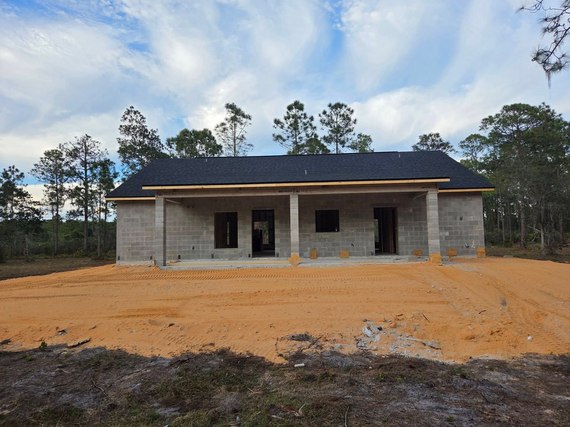 Single-story unfinished house with a dark roof on a sandy lot under a cloudy sky.