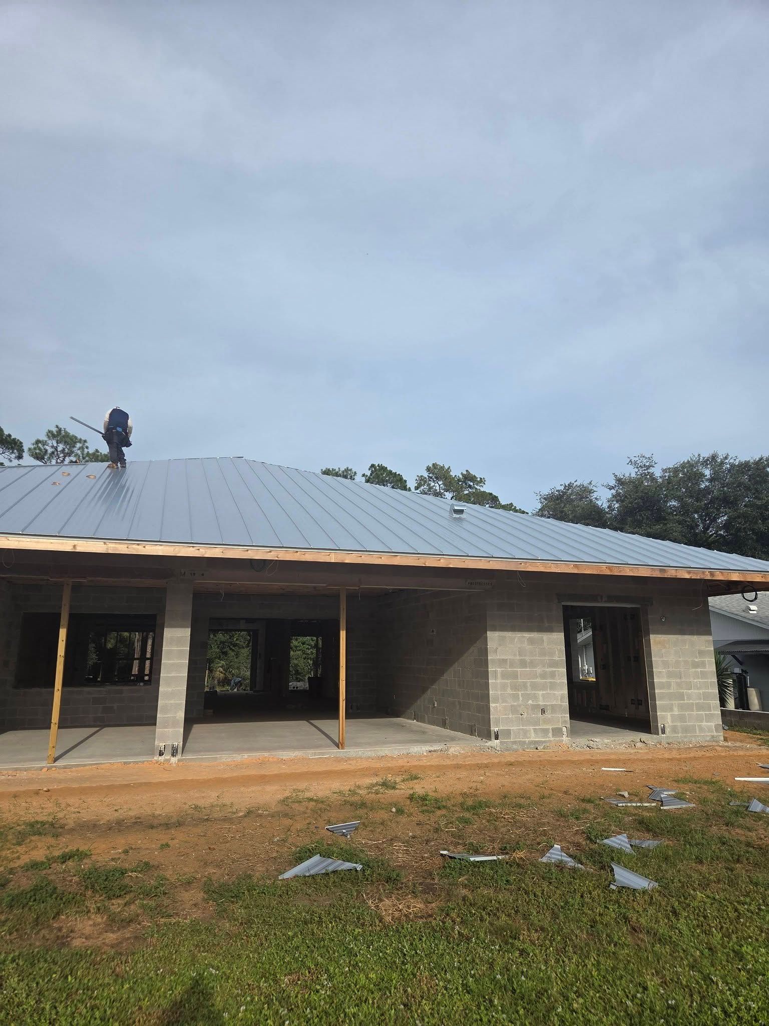 Single-story building with a metal roof under construction, with workers on the roof and dirt in front.