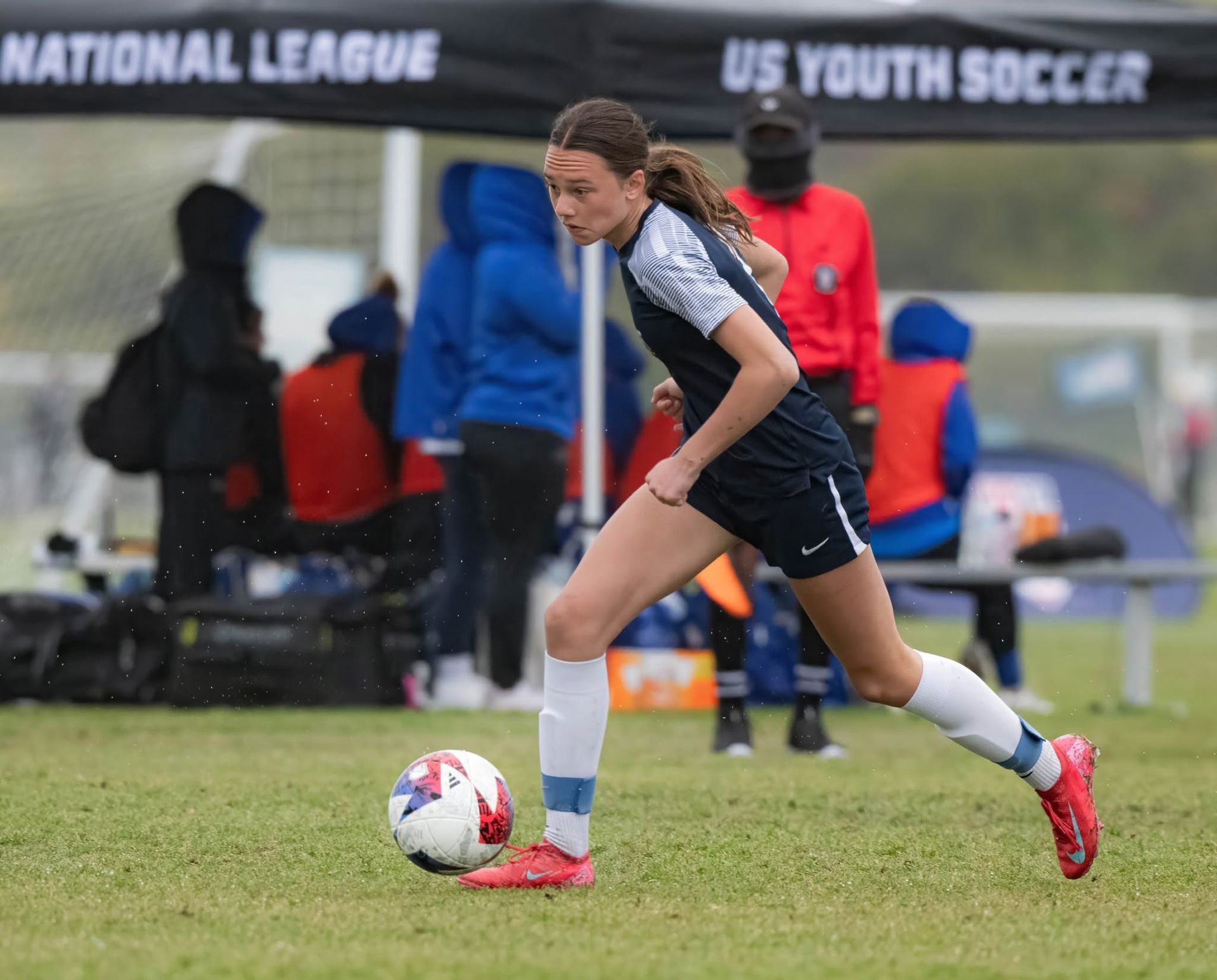 Youth soccer player in navy and white dribbles a ball on a rainy field during a match.