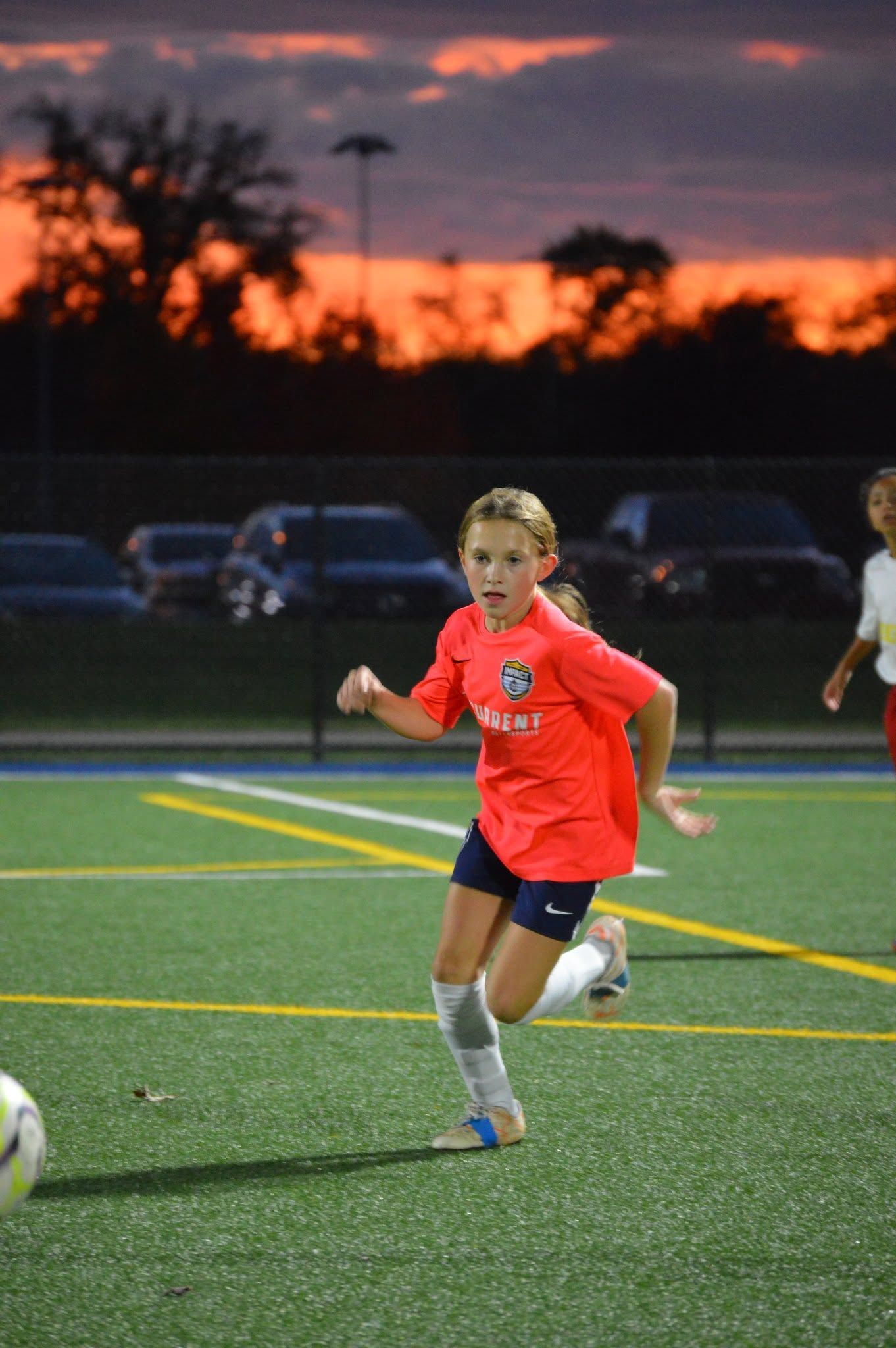 Youth soccer player in orange jersey dribbling on lit field at sunset