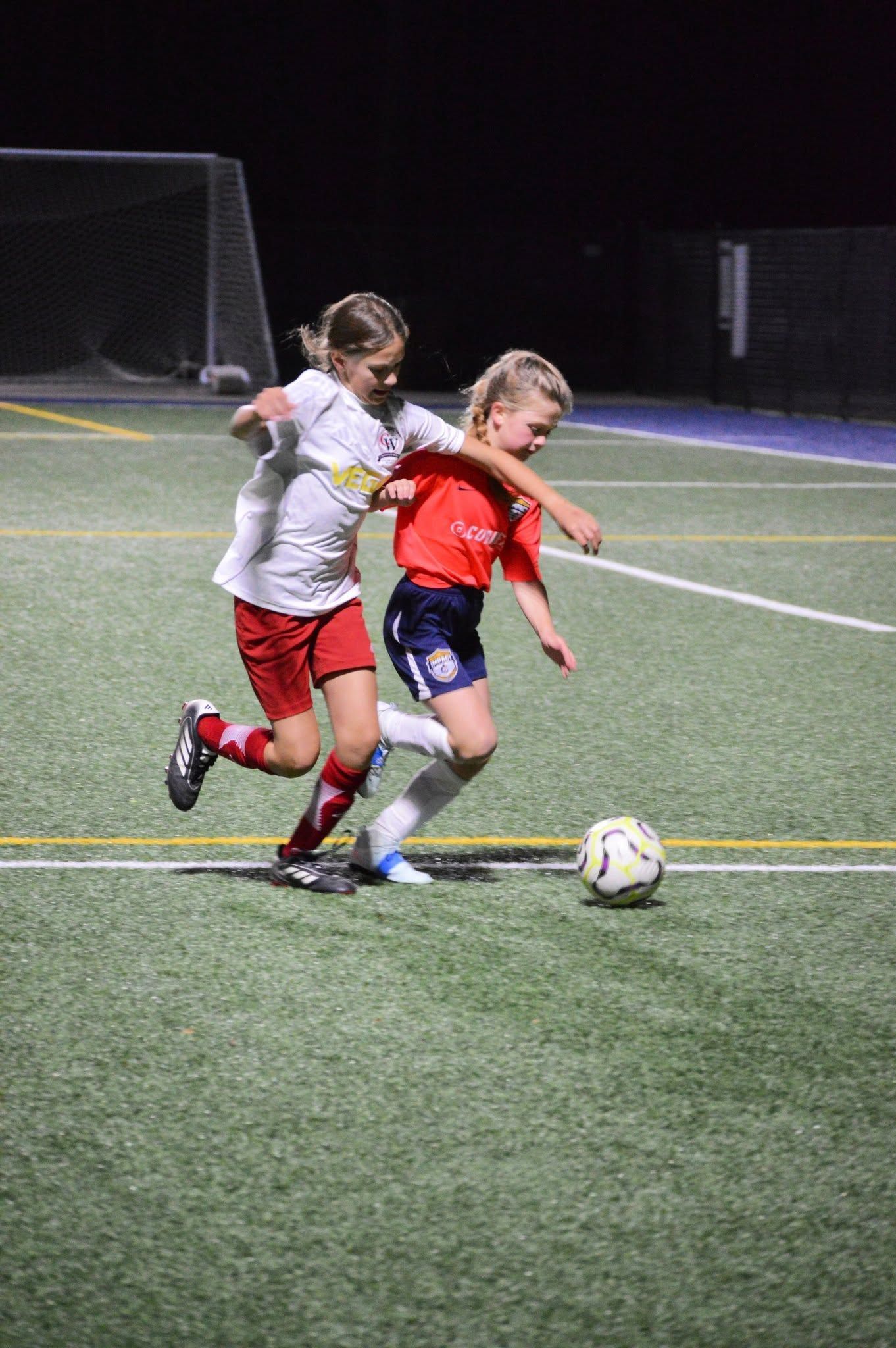 Two soccer players contest the ball on a lit field at night.