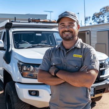 A tradesman is standing in front of a white truck with his arms crossed. There are garage doors in the background. 