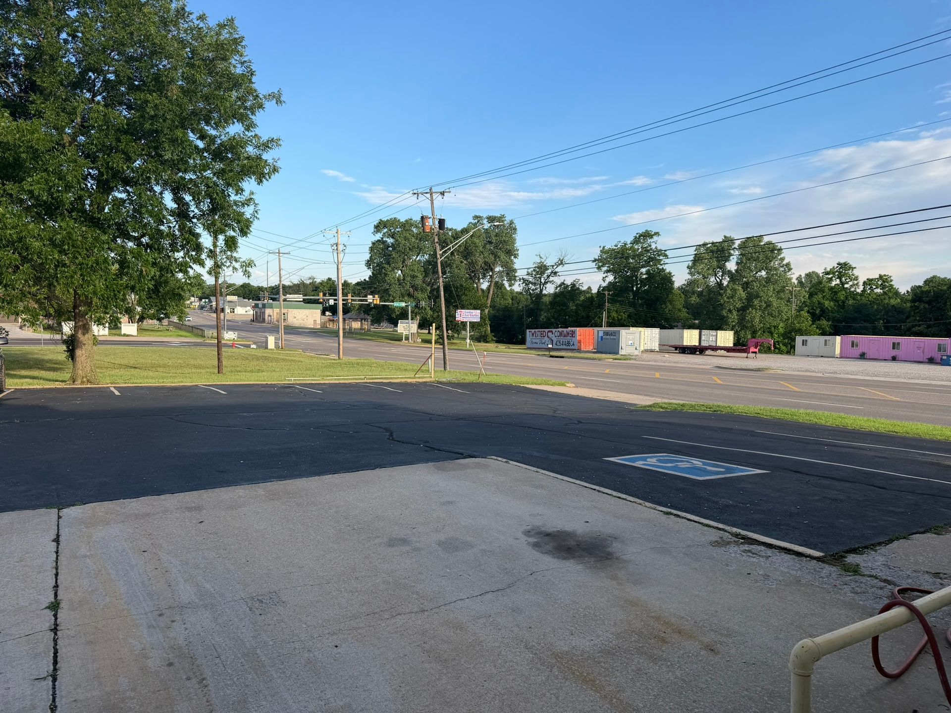 Empty parking lot beside a grassy field under a blue sky, with trees and utility poles in the background.
