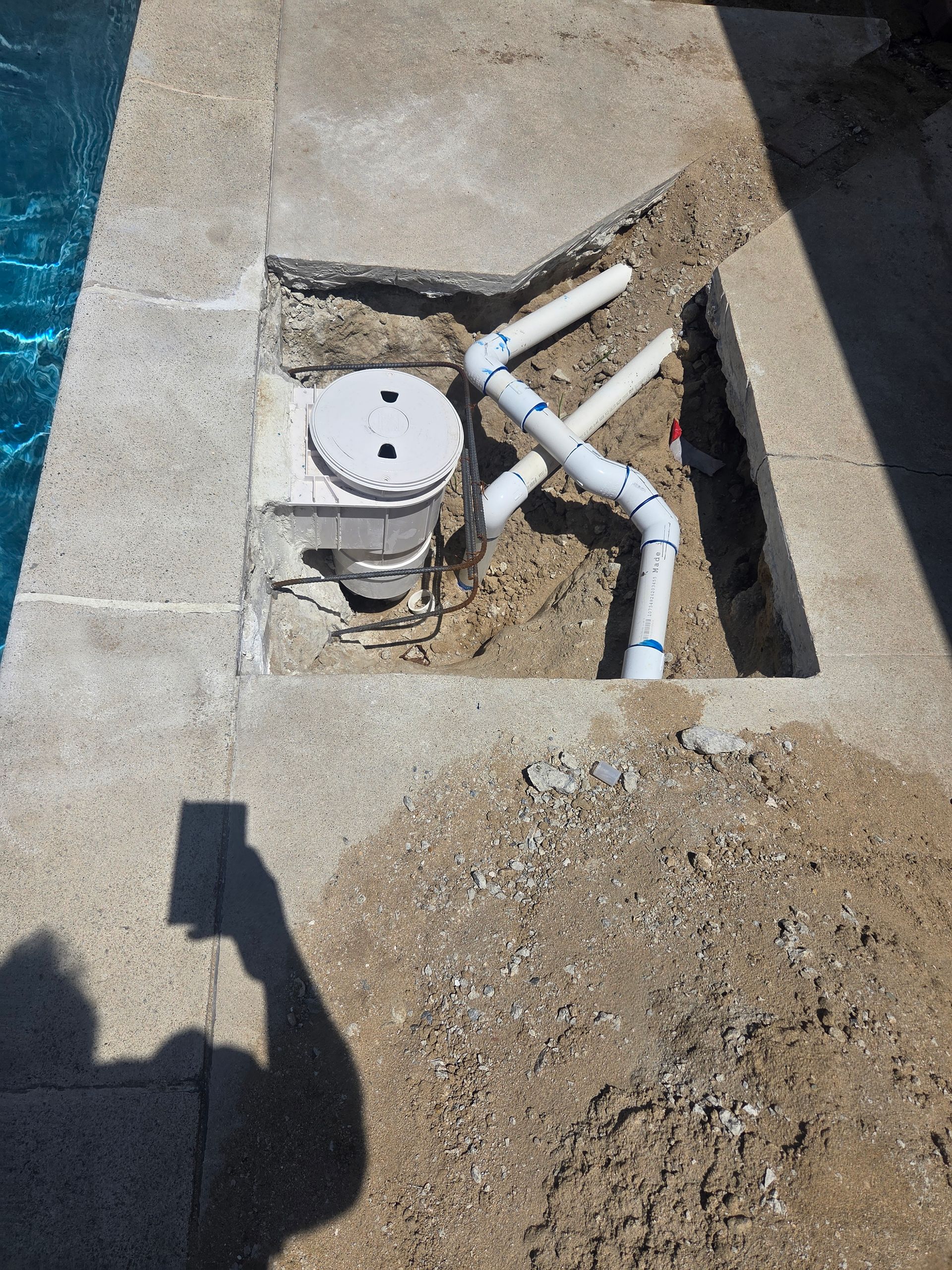 Open utility pit with white pipes and a round meter beside a pool deck.