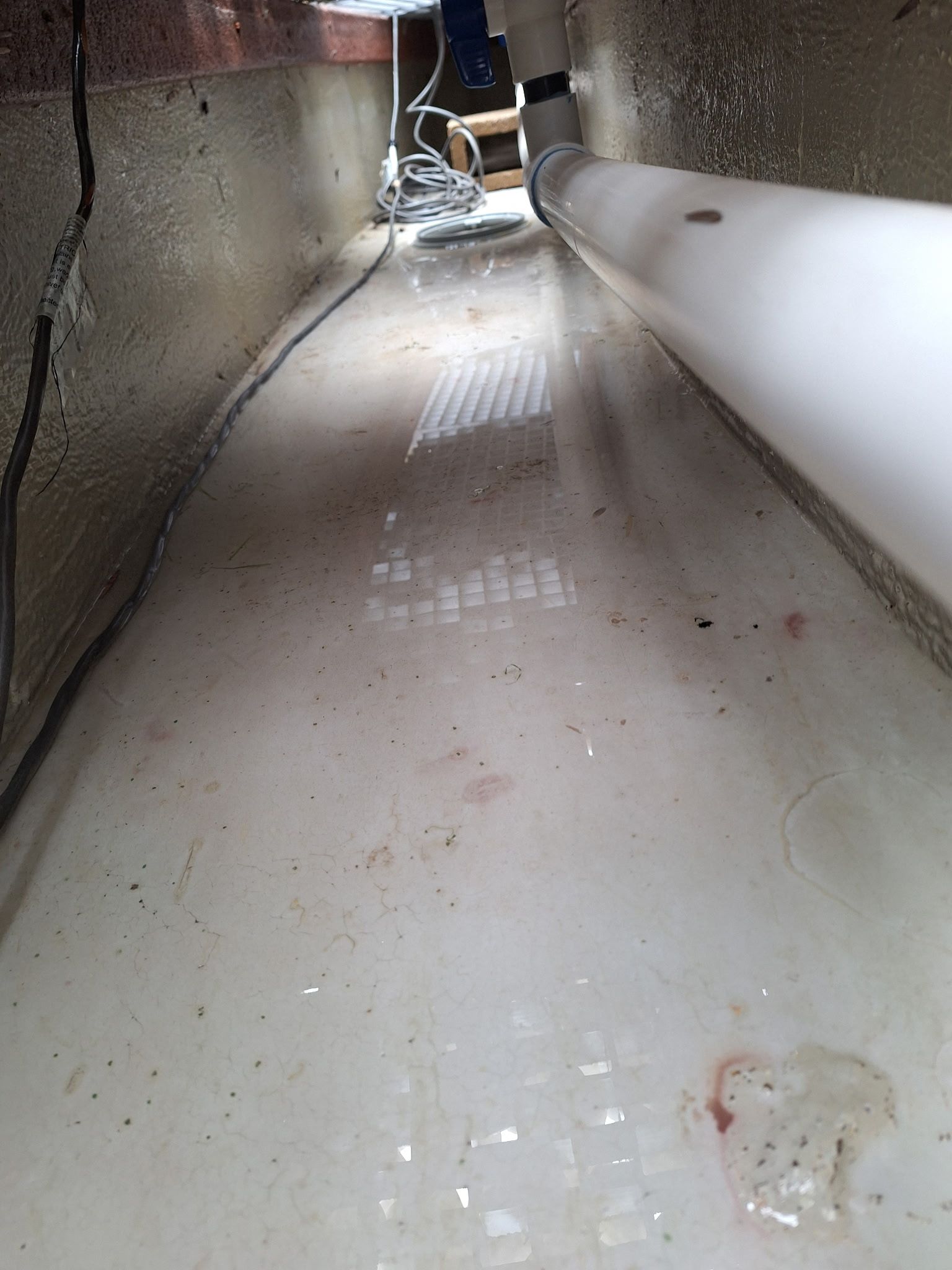 Narrow white tunnel with wet floor and debris, leading toward a small opening at the end