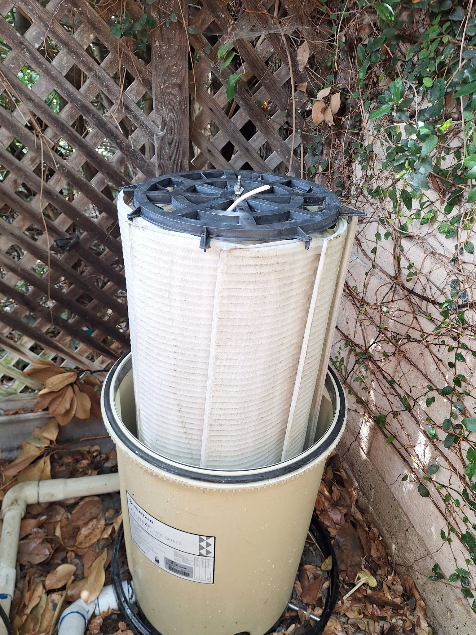 Outdoor water filter cartridge standing in a beige housing beside a lattice fence and leaves