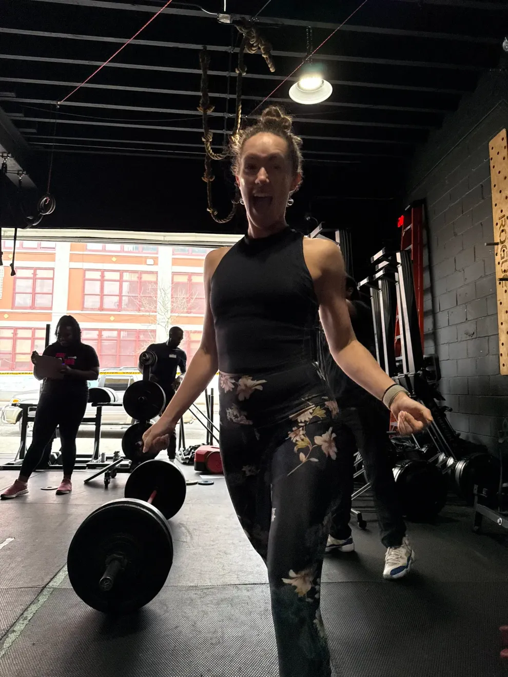 Woman lifting dumbbells at Dean CrossFit, gym in Brooklyn, NY, with large windows and another person in the background.