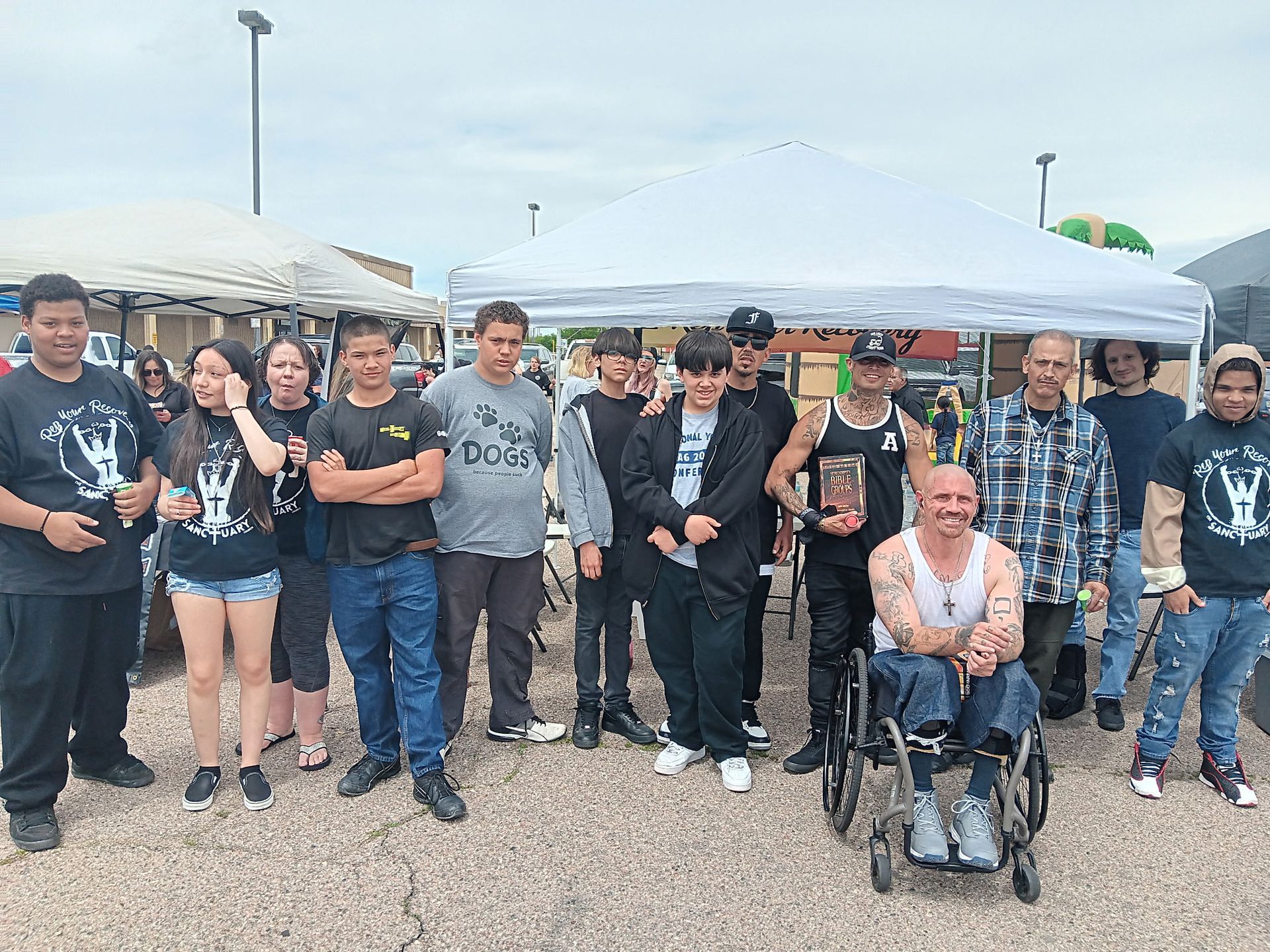 Group of people posing outdoors at a car show, with classic cars and a person in a wheelchair in front.