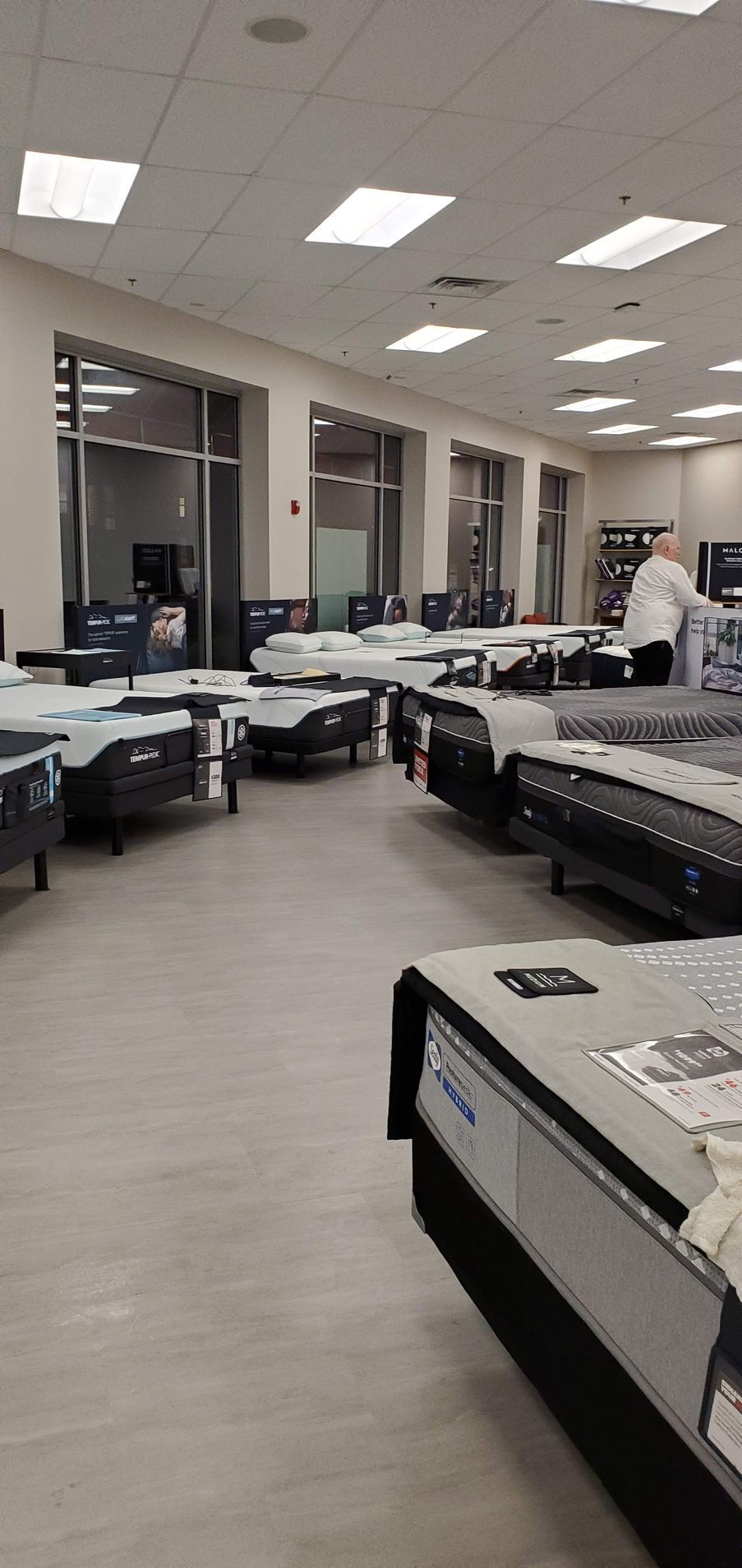 A showroom displays many mattresses on the floor, lit by overhead lights.