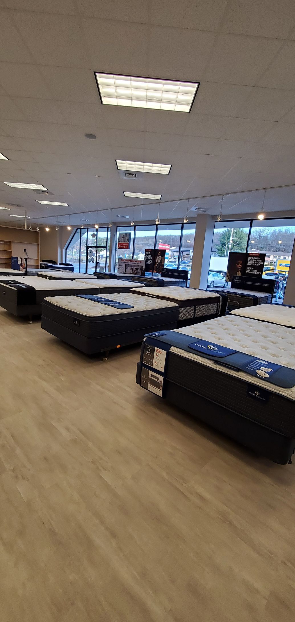 Mattress store interior with rows of displayed mattresses, overhead lighting, and large windows.
