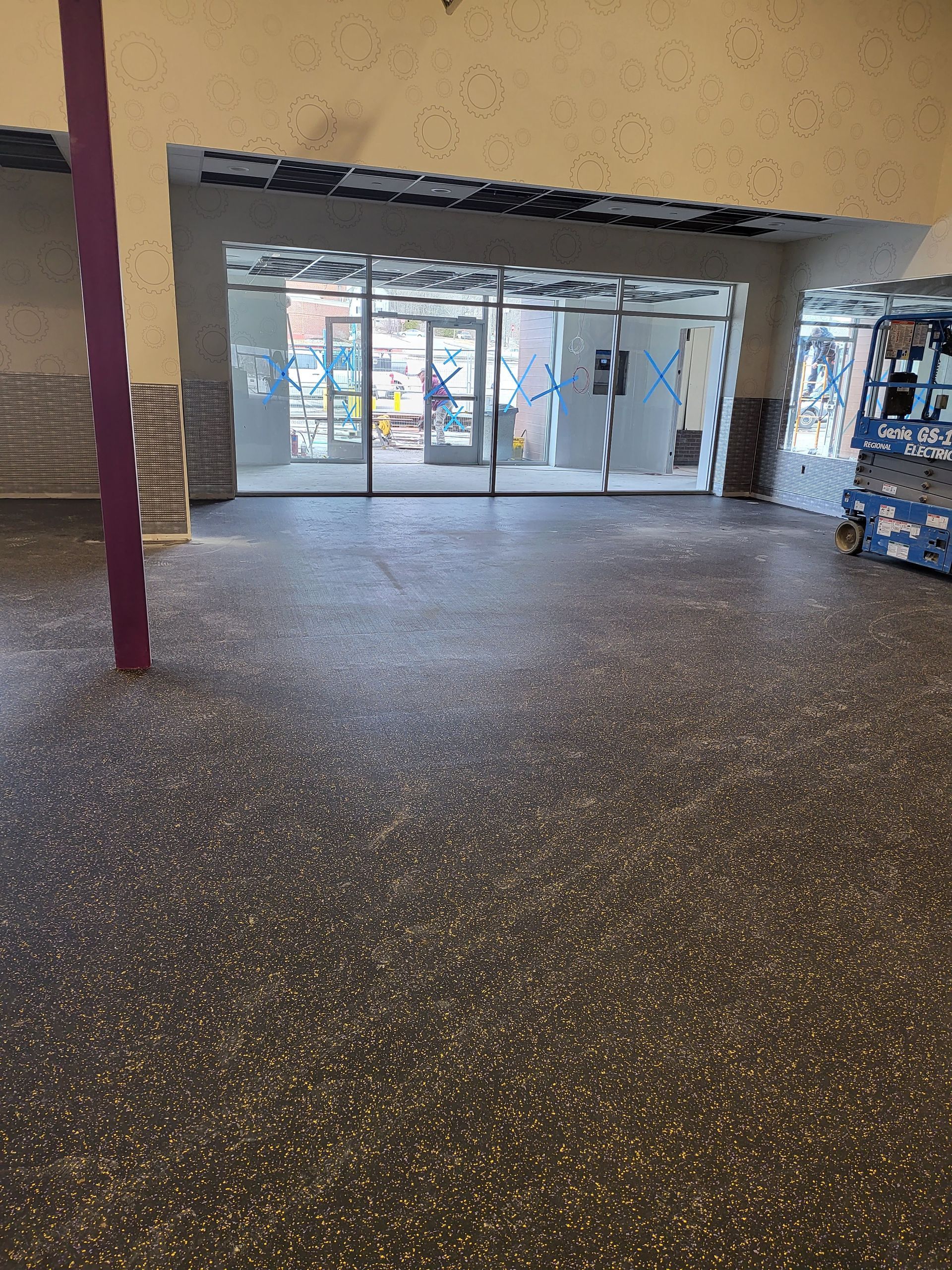 Interior view of a building's entrance with glass doors, a gray speckled floor, and a purple support beam.