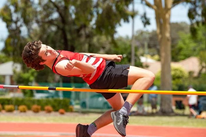 Male athlete high jumping over a yellow bar at an outdoor track meet, wearing red and black uniform.