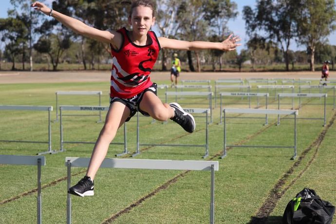 Young athlete jumps a hurdle on a green track, arms outstretched.