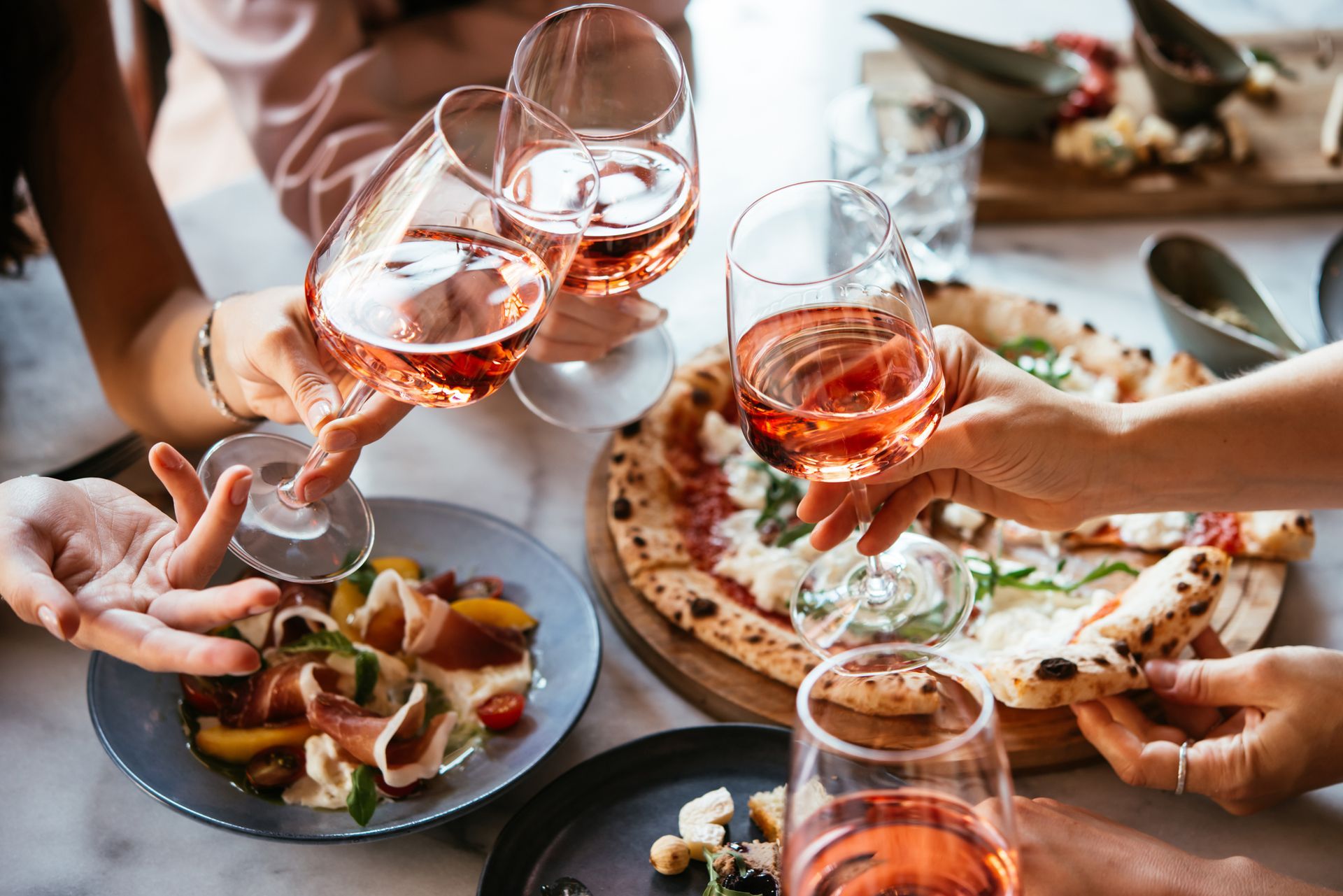 Persone che brindano con bicchieri di vino rosato su un tavolo con del cibo, tra cui pizza e insalata.