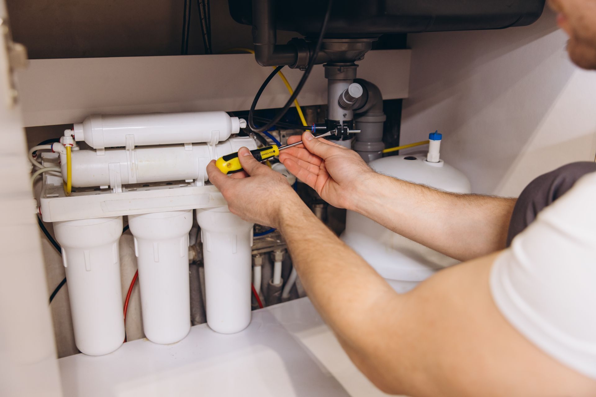 Person working on water filtration system under a sink, using a screwdriver.