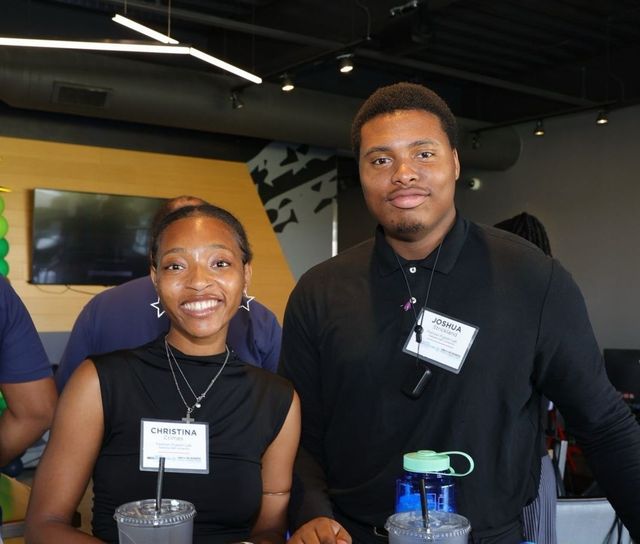 Two young Black adults smile at the camera, standing behind a counter with drinks. They are indoors with a modern design.