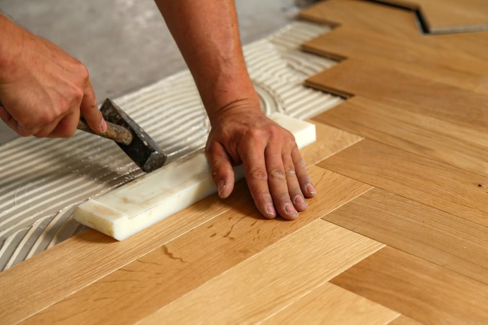 A Person Is Applying Glue to A Wooden Floor with A Hammer — John Chisholm Flooring in Atherton, QLD