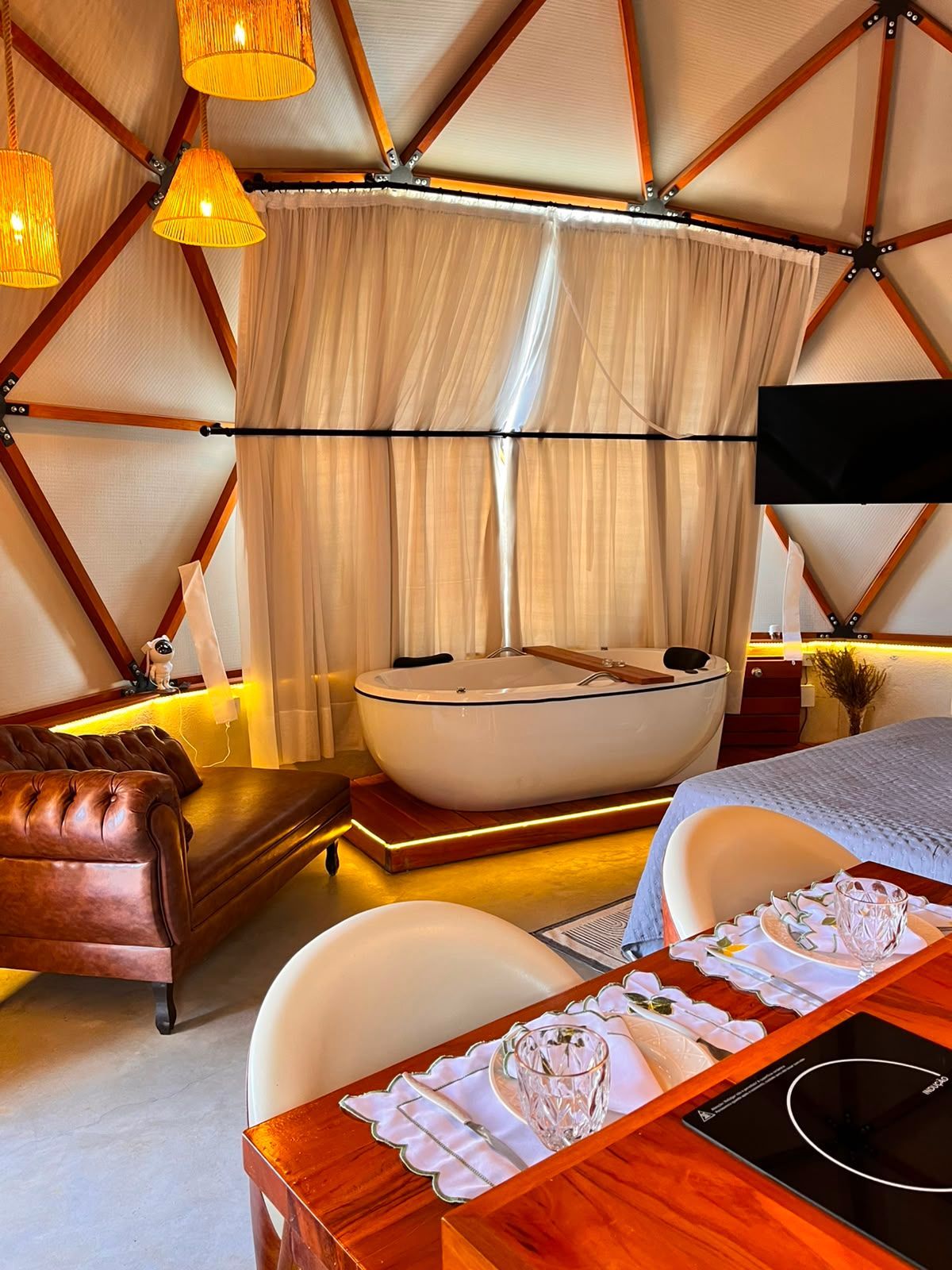 Suíte moderna de luxo em tenda com cama, banheira, poltrona de couro e iluminação âmbar aconchegante.