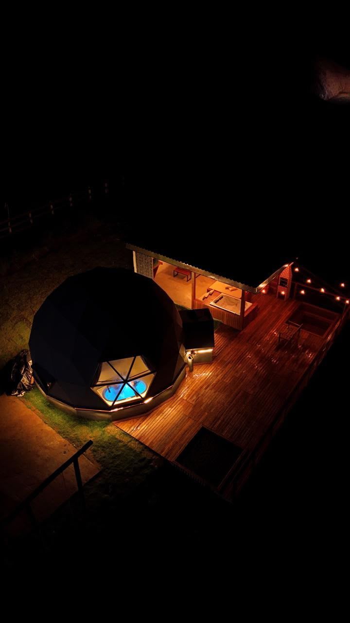 Vista aérea noturna de uma cúpula geodésica brilhante e um deck de madeira cercados pela escuridão.