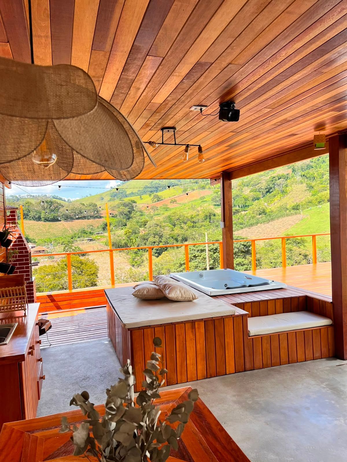 Deck de madeira ao ar livre com uma espreguiçadeira almofadada com vista para colinas e montanhas verdejantes.