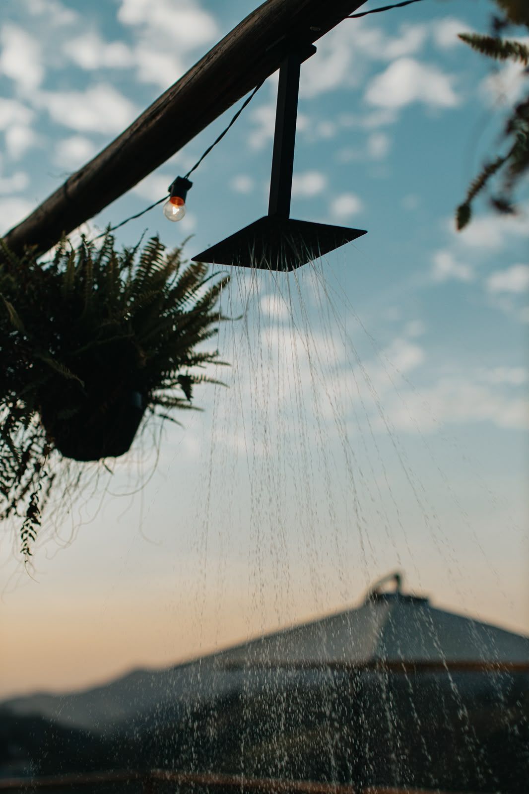 Chuveiro externo jorrando água ao pôr do sol, com planta suspensa e silhueta do telhado contra um céu nublado.