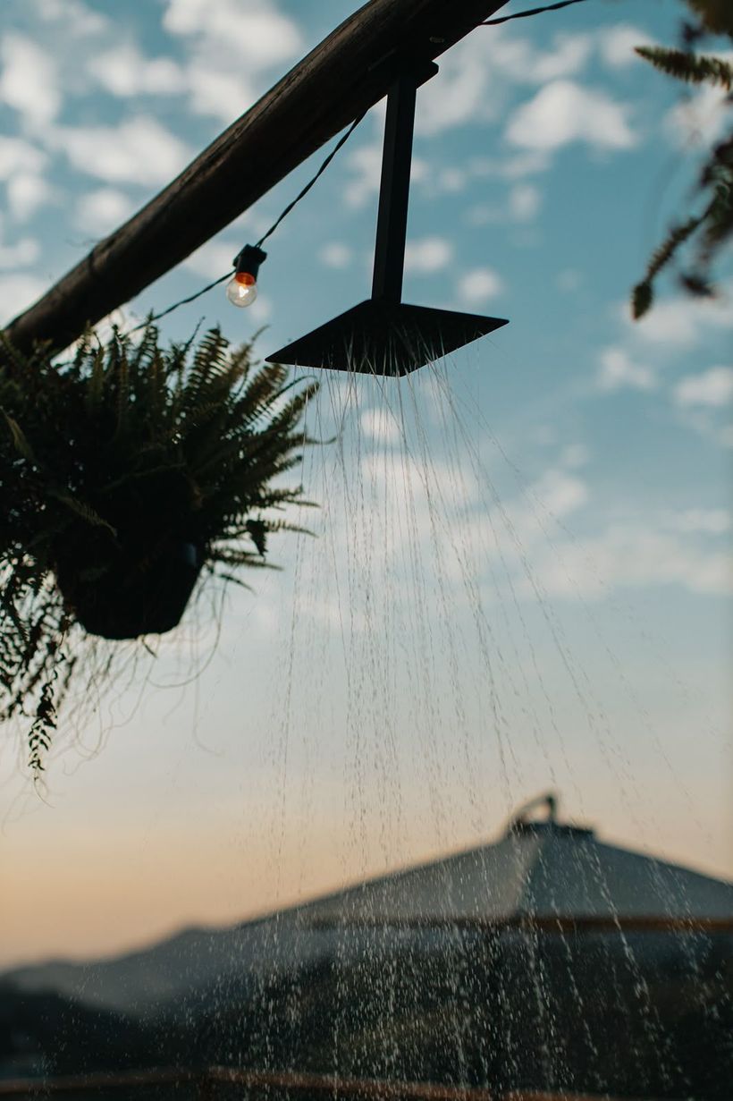 Chuveiro externo jorrando água ao pôr do sol, com uma planta suspensa e o telhado de uma casa ao fundo.