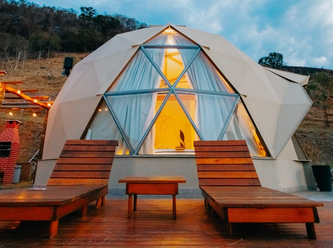 Domo geodésico para glamping com interior iluminado, cortinas e espreguiçadeiras de madeira ao entardecer.
