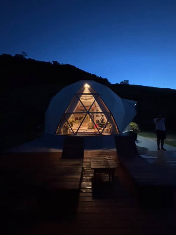Domo geodésico iluminado à noite sobre um deck de madeira, com uma pessoa em pé nas proximidades sob um céu azul.