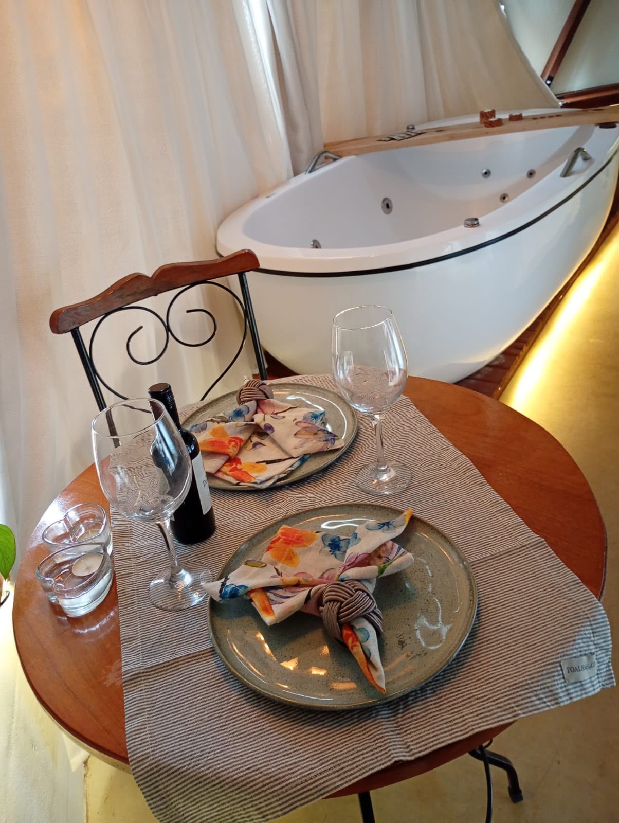 Mesa romântica posta ao lado de uma banheira branca, com taças de vinho, pratos e iluminação suave.