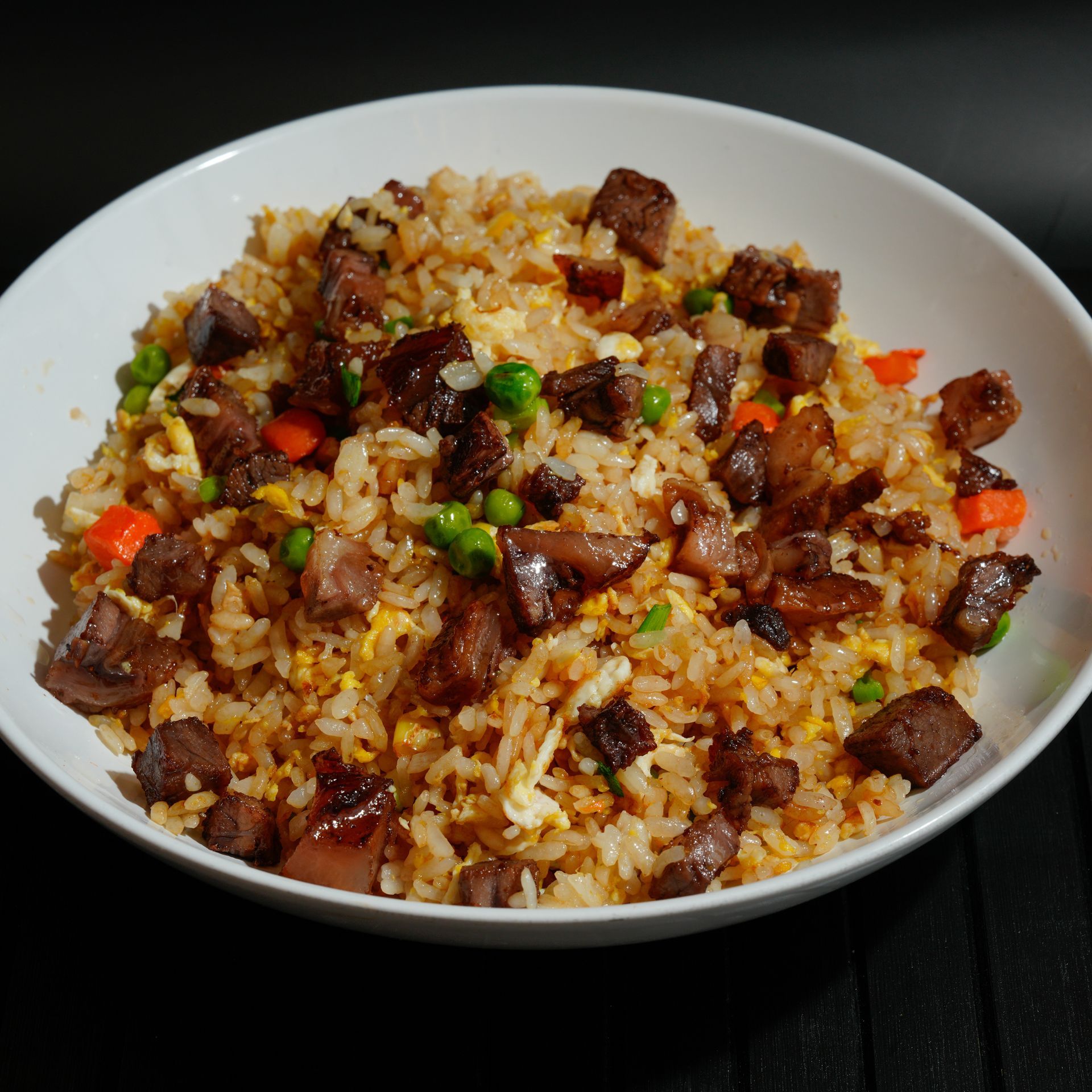 White bowl of fried rice with beef, peas, and diced vegetables on a dark table