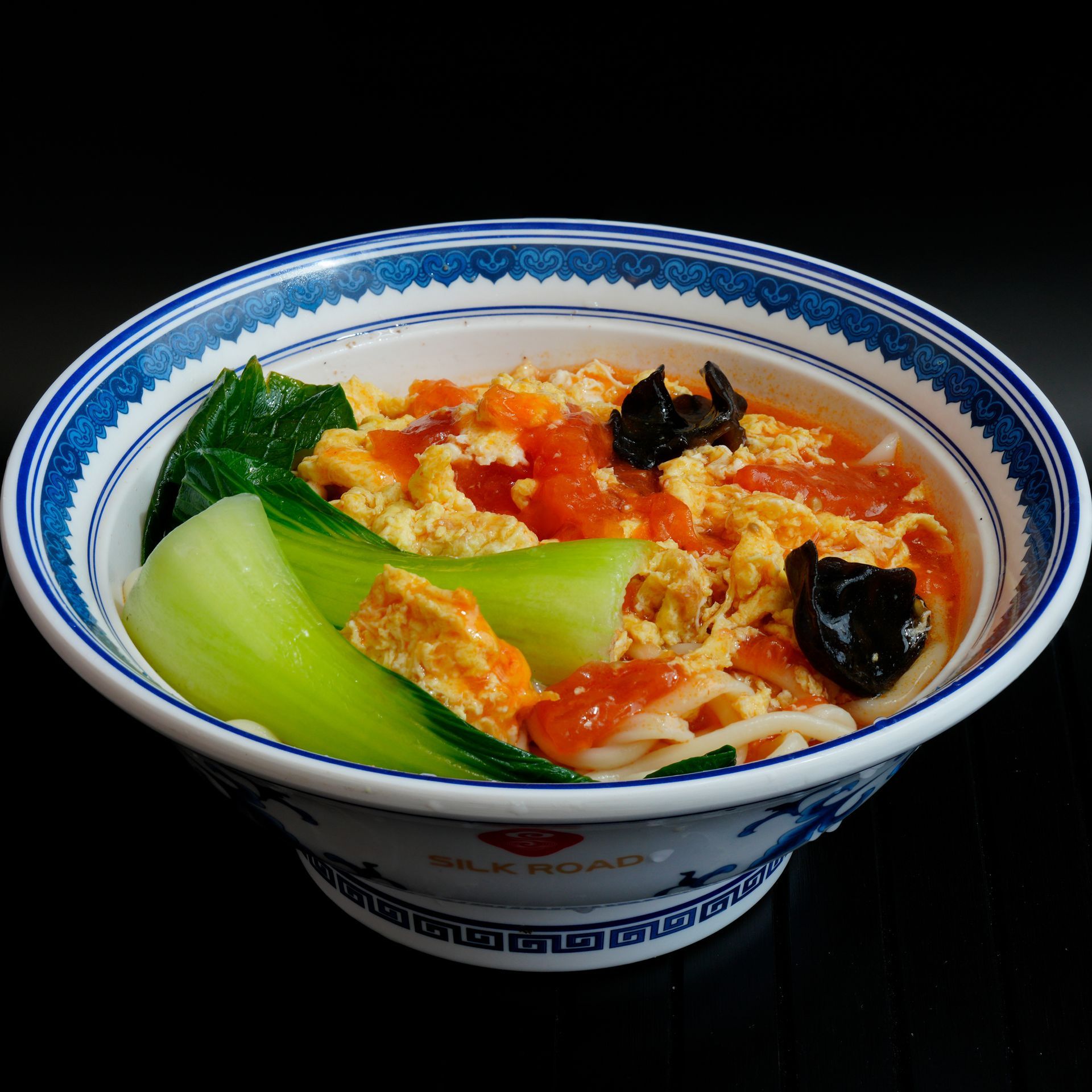 Blue-and-white bowl of noodle soup with bok choy, egg, tomato, and mushrooms on a black background