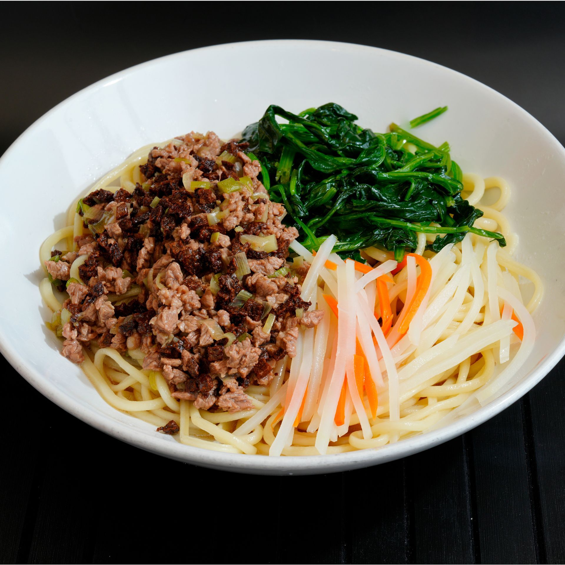 Bowl of noodles topped with minced meat, spinach, and julienned carrots on a dark table