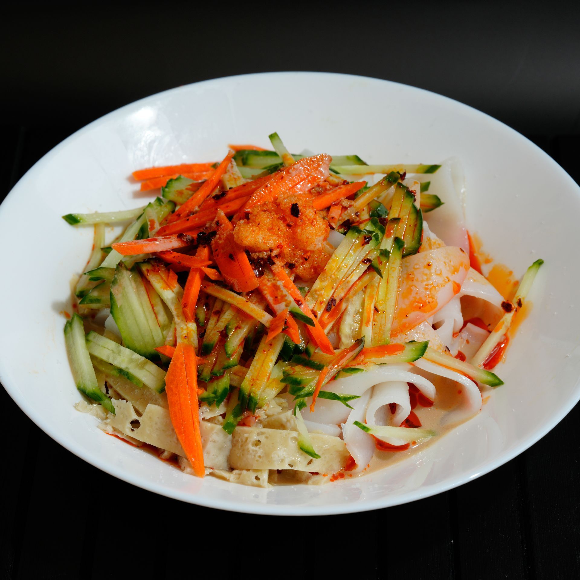 White bowl of noodle salad topped with shredded carrots, cucumber, and red chili on a dark table