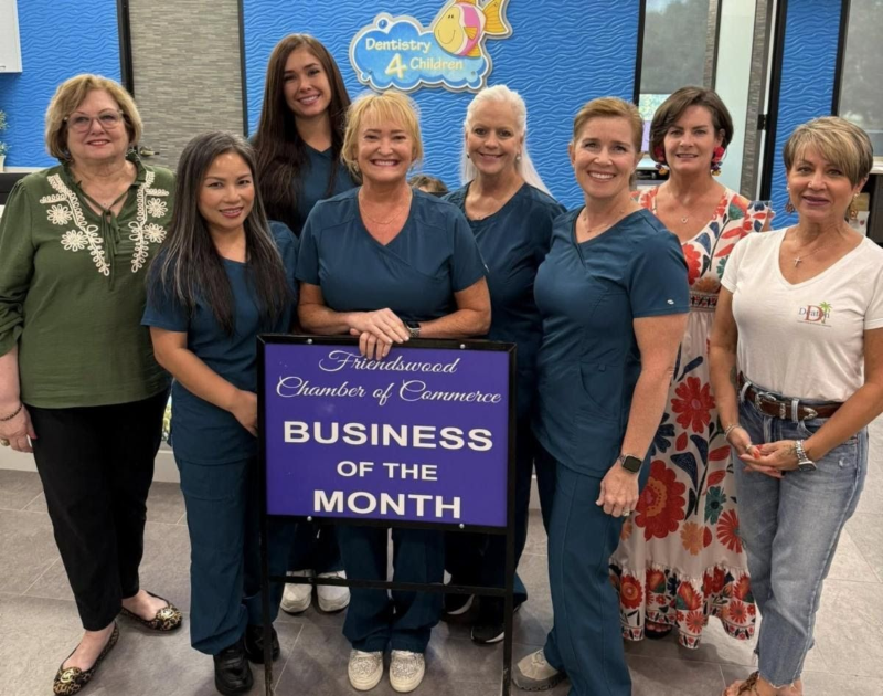 Seven people posing indoors around a “Business of the Month” sign, smiling at the camera.
