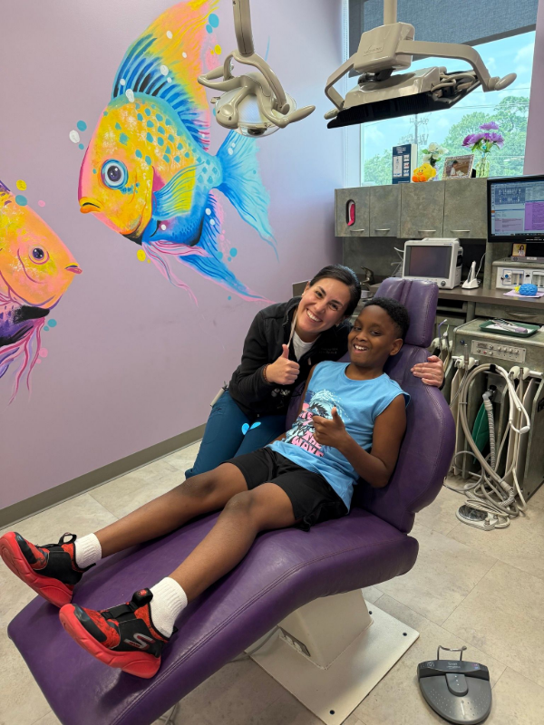 Person and child in dental chair, smiling, with colorful fish mural and dental equipment nearby.