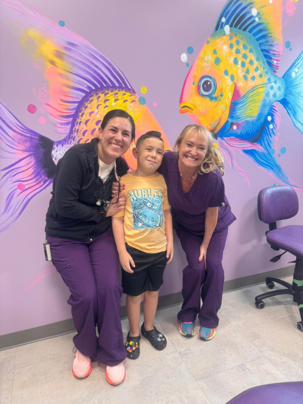 Three people smiling in front of colorful fish mural, with a chair at right.