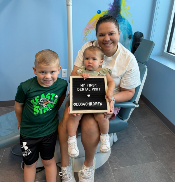 A smiling person sits in a dental chair holding a toddler who carries a sign reading 