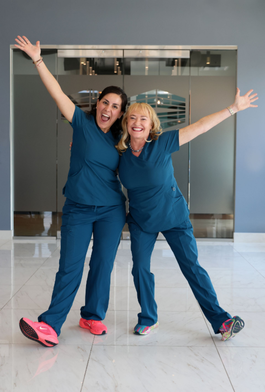 Two smiling individuals wearing teal scrubs pose with arms outstretched in a bright, modern lobby with a marble floor.
